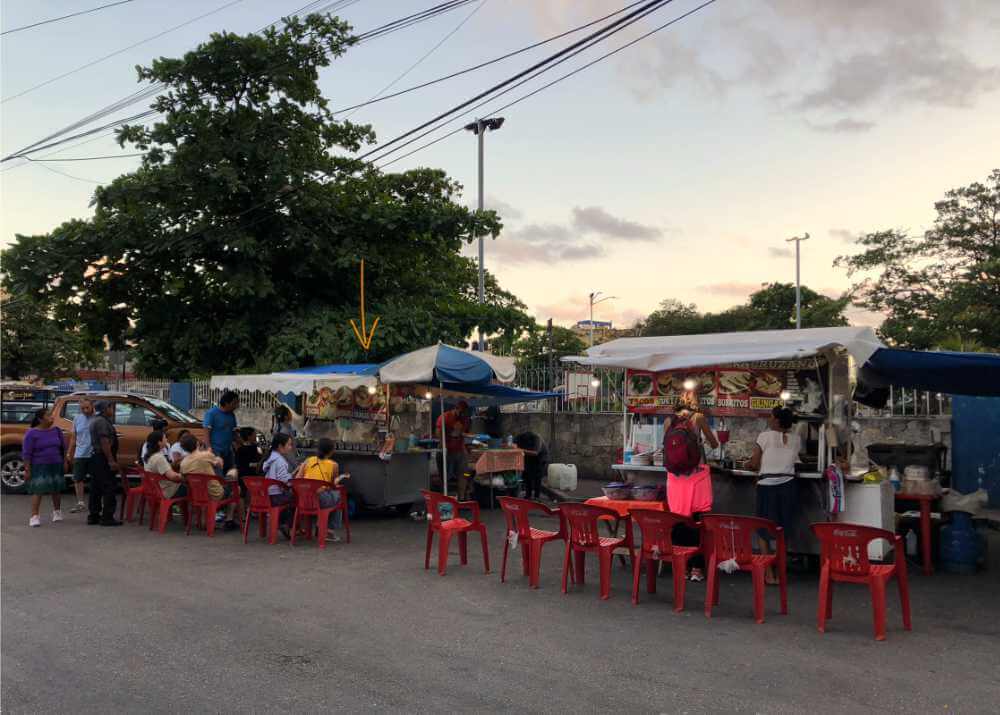 Zwei Taco-Buden am Straßenrand in Playa del Carmen. Davor stehen einige rote Plastikstühle mit einigen Gästen. Links im Hintergrund ist ein großer Baum zu sehen.