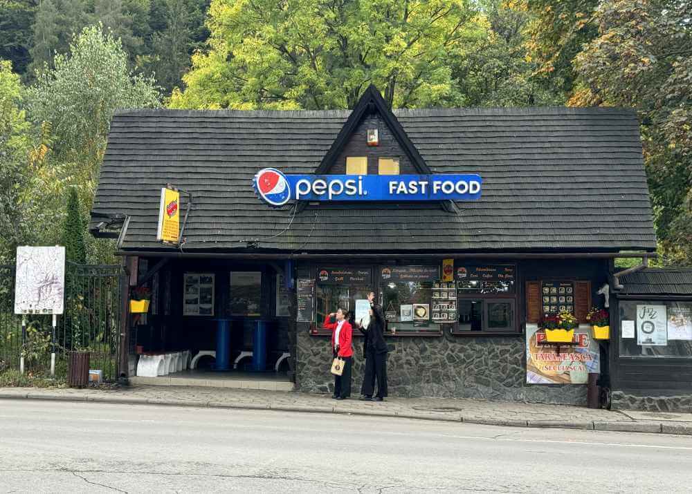 Bushaltestelle in Bran, um den Bus zurück nach Braşov zu nehmen: Ein kleines dunkles Häuschen, an dem mitten auf dem Dach ein blaues Pepsi-Schild hängt. Rechts in dem Haus ist eine Fast Food-Bude, links sind Sitzgelegenheiten. Im Hintergrund ist Wald zu sehen.