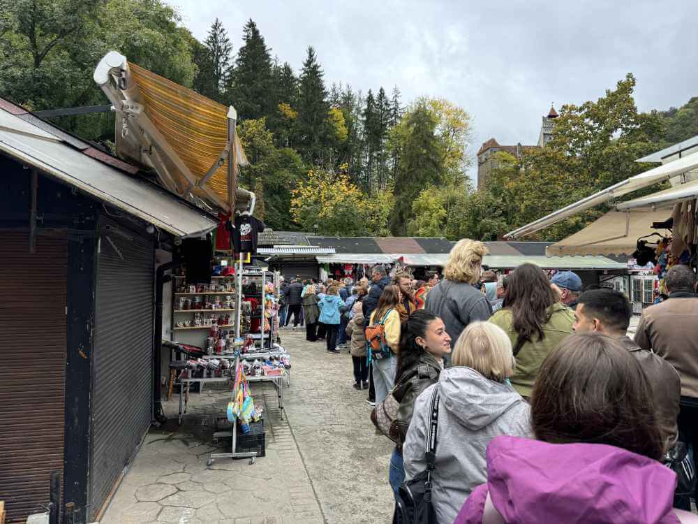 Zu sehen ist eine lange Warteschlange vor dem Eintritt zu Schloss Bran, rechts und links Souvenirbuden. Hinter dem Eingangsbereich beginnt ein Wald, links oben ist Schloss Bran zu erkennen. Das Wetter ist grau.